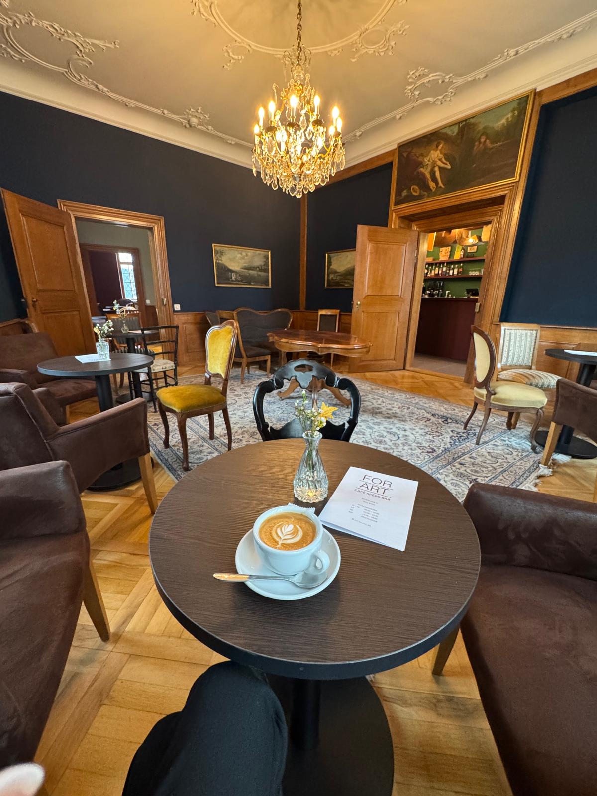 Salon der FOR ART Café Bar mit Kronleuchter, Stuckdecke und Cappuccino auf dem Tisch